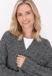 Cardigan gri, texturat, cu o croială lejeră și mâneci largi. Prezintă un design încrucișat și un material moale, pufos, cu accente în relief sub formă de bulgări.