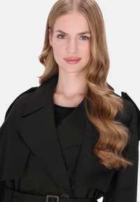 Fekete ballonkabát nagy, hajtókás gallérral és öves derékrésszel, sima textúrával. A modellnek hosszú, hullámos haja van, és semleges színű felsőt visel alatta.