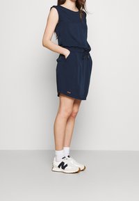 Marinblå ärmlös klänning med dragsko i midjan, sidofickor och en liten logodetalj vid fållen. Parat med vita sneakers och strumpor.
