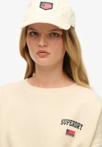 Sudadera de algodón color beige con el logo "SUPERDRY", combinada con una gorra de béisbol color crema que presenta un parche bordado de la bandera americana.
