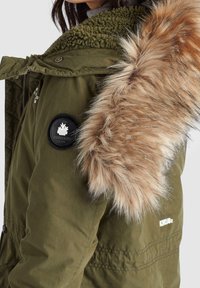 Femme portant une veste d'hiver vert olive avec un col vert duveteux et une large bordure en fausse fourrure marron clair sur la capuche, montrant les détails de côté et de dos.