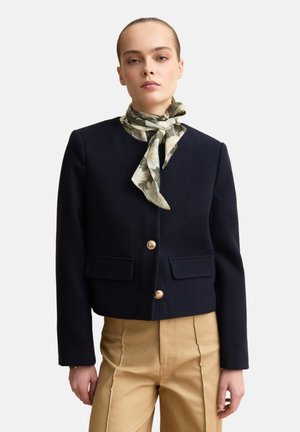 Woman wearing a black buttoned jacket, beige high-waisted pants, and a green floral patterned scarf tied around her neck.