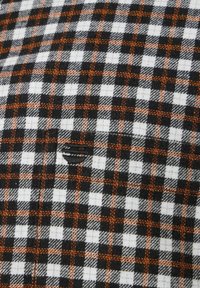 Schwarz-weiß-oranges Karostoff mit weicher Textur. Enthält kleine Quadrate und einen sichtbaren Riss im Stoff, der das Detail verstärkt.