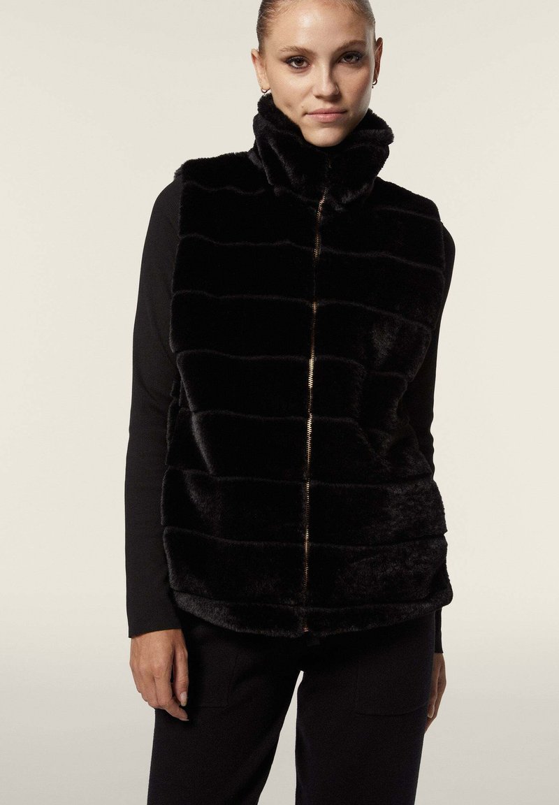 Mustat tekoturkisliivi, jossa on korkea kaulus, vetoketjulaskeuma ja vaakasuuntainen raidoitus. Pehmeä pinta ja muotoiltu istuvuus. Päällä musta pitkähihaisen paita.