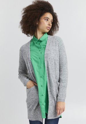 Cardigan en maille grise à ouverture frontale, à manches longues et avec poches, superposé à une chemise verte à boutons, sur fond clair.