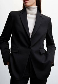 Musta räätälöity bleiseri, jossa on yksi nappikiinnitys, kaksi etutaskua ja terävät liepeet. Päällä vaalea poolopaita.