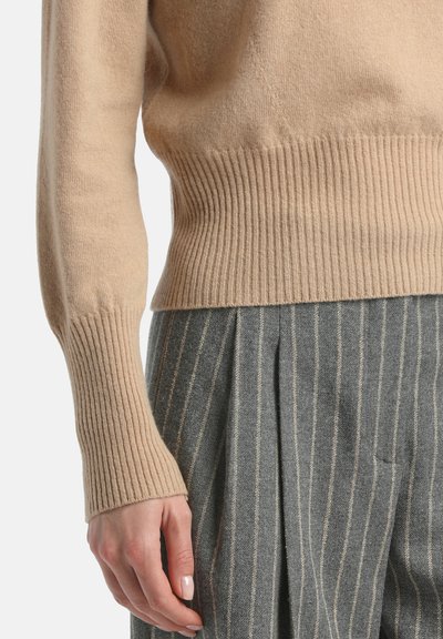 Kreminės spalvos kašmyro megztinis su riebiais rankogaliais, derinamas su pilkomis raukšlėtomis kelnėmis, turinčiomis vertikalių juostelių ir švelnų tekstūrą.