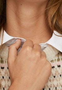 Camicia bianca con colletto parzialmente abbottonato, con una mano che stringe il colletto. Sullo sfondo si vede un indumento lavorato a maglia con motivi pastello.