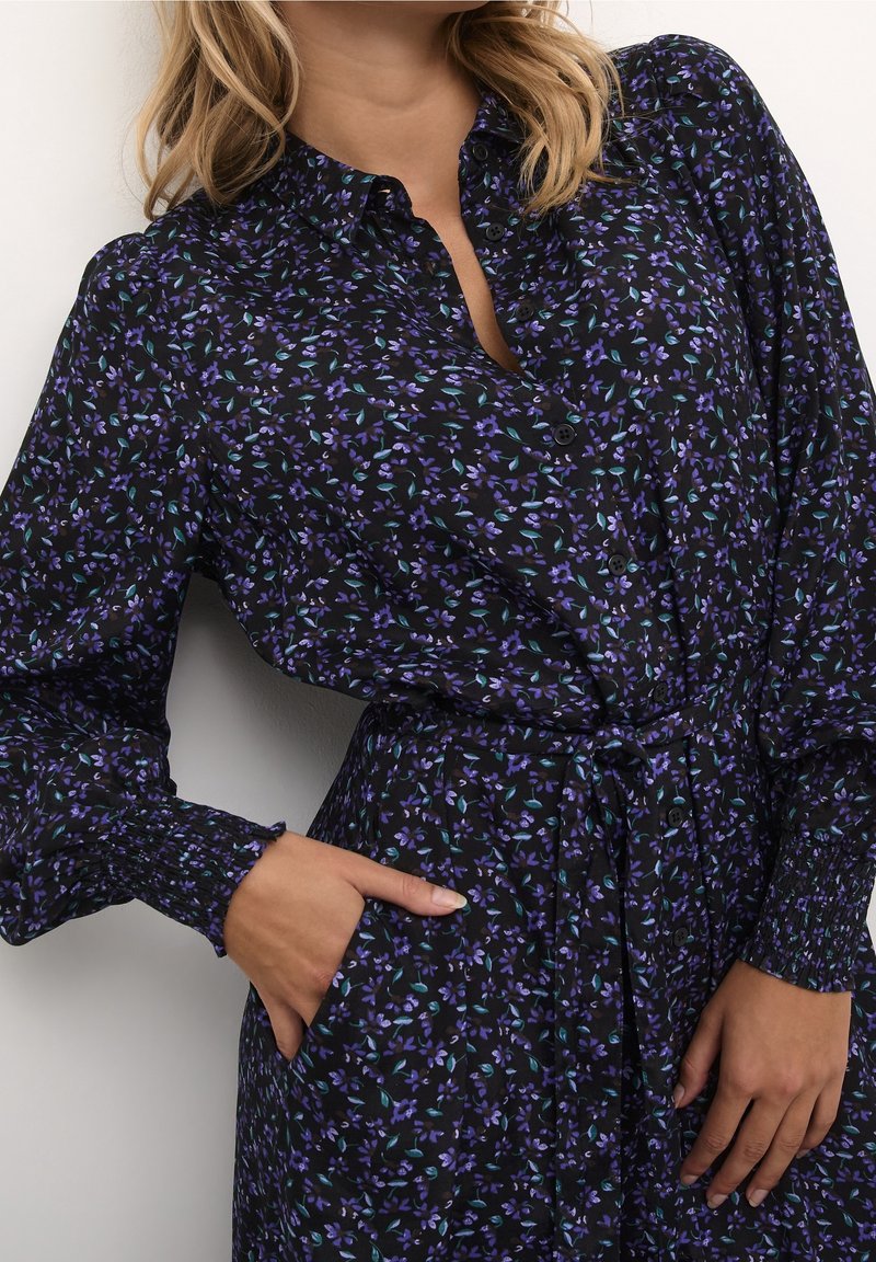 Femme portant une robe noire avec un imprimé floral bleu et violet, manches longues bouffantes, poignets froncés, boutonnage devant et ceinture nouée à la taille.