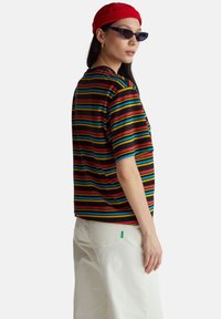 Proužkovaná krátká košile s krátkými rukávy v černé, červené, modré, žluté a zelené barvě. Má volnější střih a kapsu na levé straně hrudi. Kombinována s bílými kalhotami.