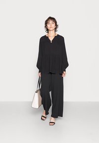 Blusa preta, oversized, com decote em V e detalhes franzidos, combinada com calças pretas de perna larga. Acessorizada com uma mala bicolor e sandálias.