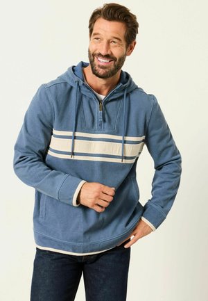 Lächelnder Mann mit blauem Kapuzenpullover mit cremefarbenen Streifen und dunkler Jeans, stehend vor einem schlichten hellen Hintergrund.