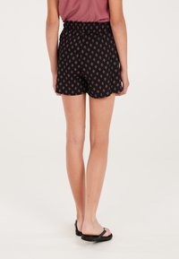 Zwarte shorts met een gatherings elastische tailleband, met een klein ruitjespatroon in het rood. Schulpzoomdetail. Draag met zwarte slippers.