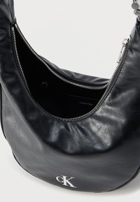 Μαύρη δερμάτινη τσάντα ώμου με ανοιχτό εν μέρει ασημένιο φερμουάρ, με λευκό λογότυπο "cK" στο μπροστινό μέρος και την επιγραφή "Calvin Klein" στο εσωτερικό επένδυσης.