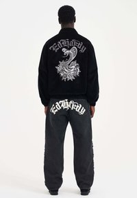 Mustat corduroy-takit, joissa on käärme- ja ruusugrafiikat selässä, yhdistettynä mustiin farkkuihin, joissa on "Ed Hardy" -logo taskuissa.