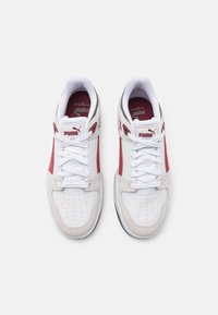 Bílé sportovní boty s červenými akcenty, s texturovanou semišovou stranou a perforovanou špičkou. Jsou viditelné šňůrky, polstrovaný jazyk a značka.