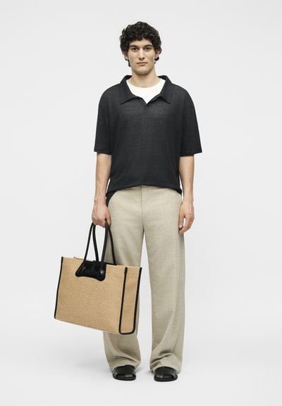 Νέος άνδρας με σγουρά μαλλιά που φοράει μαύρο κοντομάνικο πουκάμισο και μπεζ παντελόνι, κρατώντας μία μεγάλη μπεζ πλεκτή τσάντα με μαύρες λαβές.