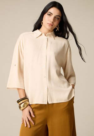 Femme aux longs cheveux foncés portant une chemise beige boutonnée, un pantalon moutarde, des boucles d’oreilles en forme d’étoile et des bracelets épais au poignet gauche.
