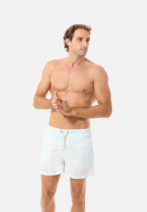 Oberkörperfreier Mann in hellblauen Badeshorts mit Kordelzug, der steht, in die Hände klatscht und nach rechts schaut vor weißem Hintergrund.