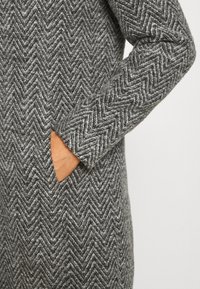 Main dans la poche d'un manteau en laine gris à motif chevrons, manche et corps visibles sur fond uni.