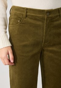 Kalhoty z korduroy v olivově zelené barvě s vertikálními žebrovanými vzory. Obsahují dvě přední kapsy a zapínání na knoflík v pase.