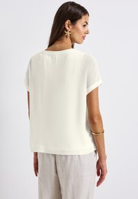 Loose-fitting, short-sleeve blouse in light cream fabric. Features a round neckline and a smooth texture. Paired with light-colored linen pants.