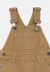 Kaki overaller med justerbare stropper, metallhardware, brystlomme og en jevn tekstur. Har en tilpasset midje og sydd sømmer.