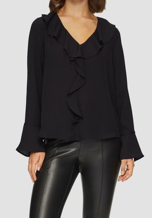 Žena v černé blůze s volánkovým výstřihem do V a manžetami, doplněná o černé kožené kalhoty, stojící před jednobarevným světlým pozadím.