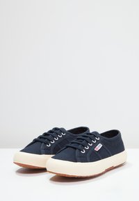 Marine lærrets sneakers med teksturert overdel, hvite snøringer og en tan gummisåle. Har en merket tagg på siden.