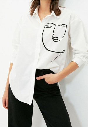 Osoba nosící bílou košili na knoflíky s abstraktním černým obličejovým uměním na přední straně, doplněná černými kalhotami a s jednou rukou v kapse.