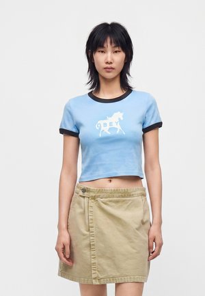 Jeune femme aux cheveux noirs portant un t-shirt court bleu clair avec une licorne blanche et une jupe portefeuille beige sur un fond uni.