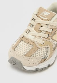 Μπεζ και κρεμ αθλητικό παπούτσι New Balance με ύφασμα από δίχτυ, σουέντ επενδύσεις, κορδόνια και γκρι ελαστική σόλα σε λευκό φόντο.