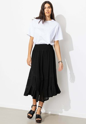Femme debout contre un mur blanc, portant une chemise blanche à manches courtes, une jupe midi noire avec des détails œillets et des sandales noires à plateforme et bout ouvert.