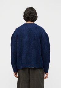 Άτομο με κοντά σκούρα μαλλιά, φορώντας χαλαρό μπλε ναυτικό πουλόβερ με υφή και σκούρο καφέ παντελόνι, εμφανίζεται από πίσω σε απλό φωτεινό φόντο.