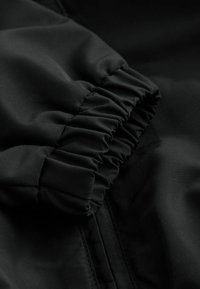 Schwarzer Stoff mit einem gerafften, elastischen Bündchen. Glatte Textur mit subtiler Glanz, mit einem Reißverschluss entlang einer Kante. Nahaufnahme.