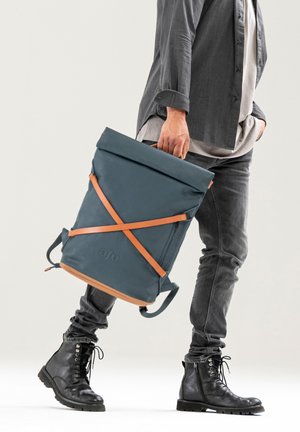 Person trägt dunkle Jeans und Stiefel, hält einen blauen Rucksack mit hellbraunen Riemen und aufrollbarem Verschluss am Griff.