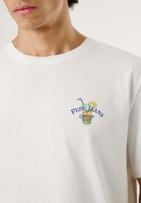 Biela bavlnená tričko s krátkymi rukávmi, ktoré má farebnú vyšitú nálepku nápoja s citrónom a textom "Pepe Jeans London S-1973" na hrudi.