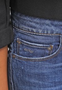 Granatowe dżinsy z małą kieszonką z przodu, metalowymi nitami i kontrastującymi beżowymi przeszyciami. Na kieszeni wyhaftowana jest marka "RAW".