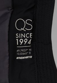 Crni tekstilni etiketa s bijelim tekstom: "QS SINCE 1994", koordinate i hashtag. Glatka tekstura s šivanim rubovima.