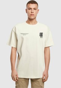 Camiseta de algodón crema con un corte relajado, que presenta un estampado gráfico negro en el pecho y texto a lo largo del lateral. Manga corta, escote redondo.
