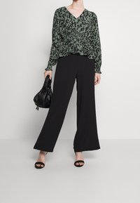 Bluse mit grünem und schwarzem Leopardenmuster, V-Ausschnitt und geraffter Taille, kombiniert mit schwarzen weit geschnittenen Hosen und schwarzen Absatzsandalen.