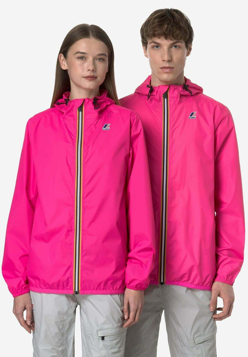 Due modelli indossano giacche con cappuccio K-Way di colore rosa acceso con cerniere a righe e pantaloni cargo grigio chiaro, in piedi fianco a fianco.