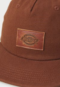 Brun hatt laget av teksturert stoff, med en rektangulær lærflekke med preget logo. Seks panelkonstruksjon med toppknapp og søm.