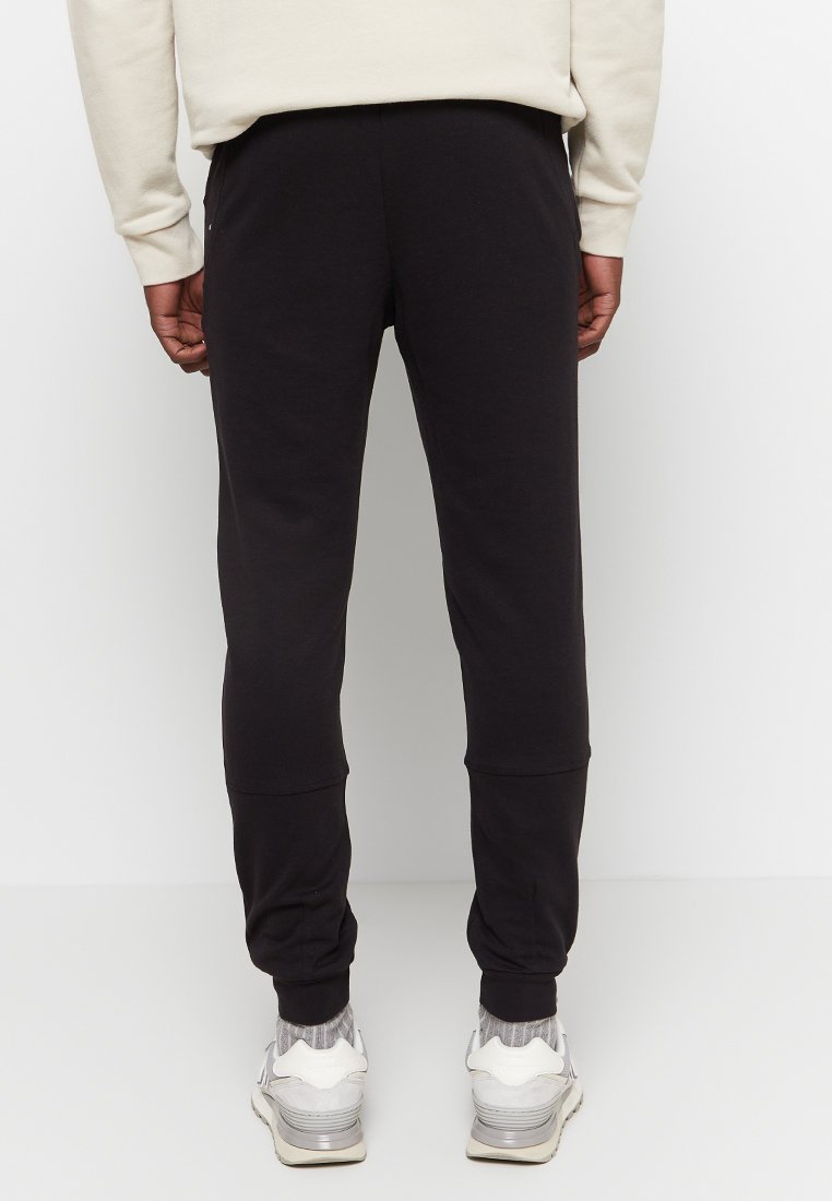 Pantalones de chándal negros hechos de una tela suave, con un diseño ajustado, puños acanalados y una textura lisa. Se llevan con zapatillas de color claro.