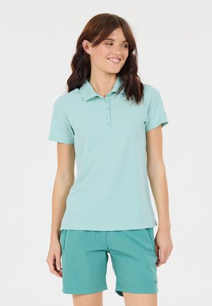 Kvinde med skulderlangt brunt hår iført en lyst turkis polo og teal shorts, stående og kigger sidelæns med et svagt smil.