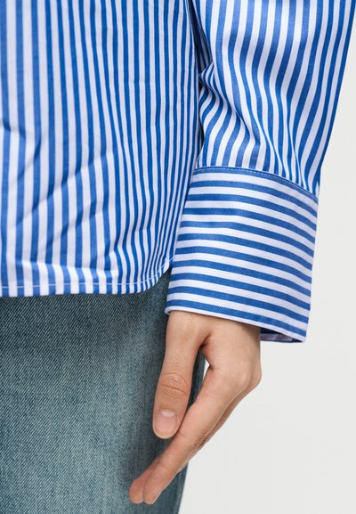 Χέρι που ακουμπά στο πλάι, φορώντας μπλε και λευκό πουκάμισο με κάθετες ρίγες μακριού μανικιού και ανοιχτόχρωμο τζιν παντελόνι.