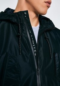 Jauna persona, tērpusies melnā kapučjakā ar uzrakstu "BIG STAR LIMITED" uz rāvējslēdzēja pārsega un pelēku apakškreklu, kas redzams pie apkakles.