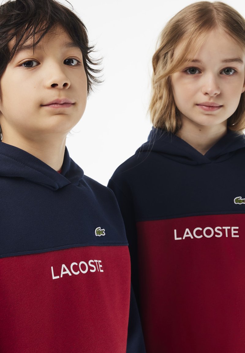 Zwei Kinder tragen passende Hoodies. Die Hoodies sind navyblau und rot, aus weichem Material gefertigt und tragen das "LACOSTE"-Logo sowie Logo-Stickerei.