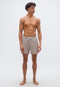 Νέος άνδρας με σγουρά μαλλιά που φοράει μπεζ και λευκά κάθετα ριγέ μαγιό, όρθιος ξυπόλητος μπροστά σε ένα απλό φωτεινό φόντο.