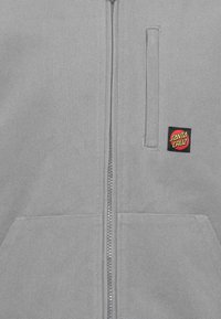 Γκρι φούτερ με μισό φερμουάρ και μπροστινές τσέπες, με μικρό κόκκινο και κίτρινο λογότυπο της Santa Cruz στην αριστερή περιοχή του στήθους.